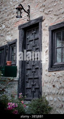 Affascinante porta in legno antica con una struttura in legno, adagiata in uno storico edificio in pietra. Scalini in pietra conducono all'ingresso, adornati da fiori vivaci. Foto Stock