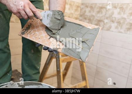 Un operaio edile applica l'adesivo di cemento su una piastrella in ceramica e lo distribuisce a fondo su tutta la superficie Foto Stock