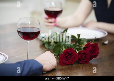 C'è un bouquet di rose rosse su un tavolo di legno. Accanto a loro ci sono piatti vuoti e due bicchieri di vino rosso. La donna ha uno degli occhiali Foto Stock