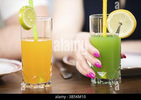 Primo piano di due bevande in bicchieri alti. Uno è arancione e l'altro è verde. Hanno entrambi le cannucce. Uno degli occhiali e' tenuto dalla h di una donna Foto Stock