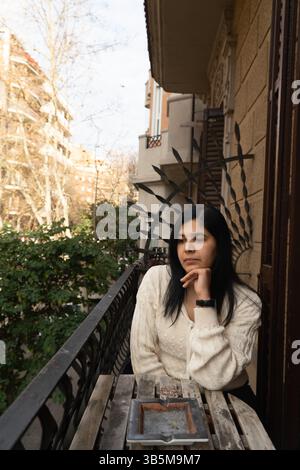 Una donna è seduta su un balcone con una sigaretta in mano. Indossa un maglione bianco e un orologio Foto Stock