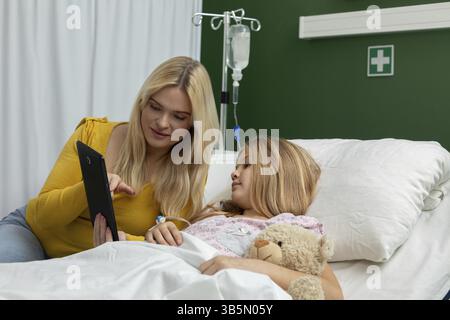 Una giovane donna conforta un paziente in un letto d'ospedale utilizzando un tablet, evidenziando la cura e la connessione Foto Stock