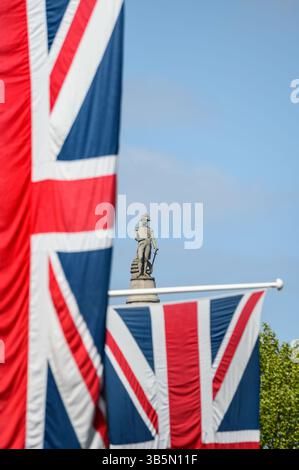 Londra, Regno Unito. La statua di Nelson in cima alla colonna di Nelson in Trafalgar Square, vista da Whitehall, dietro un albero e Union Flags appese all'esterno Go Foto Stock
