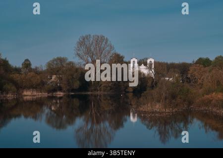 Antica chiesa cattolica in pietra bianca riflessa in acque calme con cielo blu che incombe sugli alberi scuri. Edificio storico gotico Foto Stock
