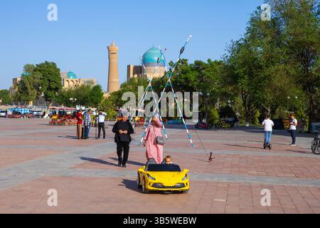 Un bambino piccolo in un'auto sportiva in miniatura gialla che ha fuori dalla Moschea di Kalan (1542) e dal minareto di Kalyan (1127) a Bukhara, Uzbekistan Foto Stock