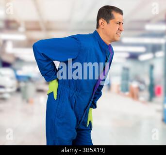 Bell'uomo di mezza età che indossa l'uniforme blu meccanico soffre di mal di schiena, toccare la schiena con la mano, dolore muscolare in officina Foto Stock