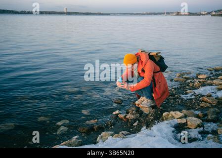 Un giovane uomo con un cappotto arancione e un berretto giallo accovacciato su una costa rocciosa parzialmente coperta dalla neve mentre scatta foto dell'acqua con il suo smartphone durante una fredda giornata invernale Foto Stock