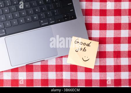Una nota adesiva gialla che mostra un buon lavoro e una faccia sorridente poggia sulla tastiera di un notebook accanto a una tovaglia a scacchi rossa e bianca, a simboleggiare i positi Foto Stock