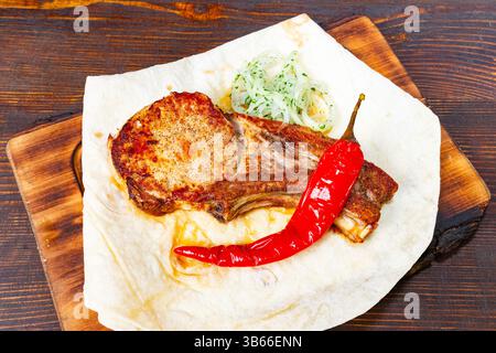 Una costoletta di maiale perfettamente cotta poggia su una tavola di legno, accompagnata da una fresca guarnizione di cipolla verde e un peperoncino rosso brillante, che mostra un appetizi Foto Stock