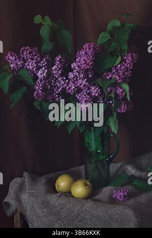 Natura morta con bouquet lilla e mele, vista a livello degli occhi. La luce soffusa della sera illumina delicatamente un lussureggiante bouquet lilla in una caraffa di vetro verde con mele. Foto Stock