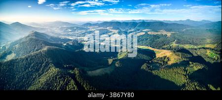 Vista aerea panoramica di un vasto paesaggio caratterizzato da colline ondulate, fitte foreste e insediamenti rurali sparsi sotto il cielo azzurro limpido con nuvole. Scena suggestiva e pittoresca. Foto Stock