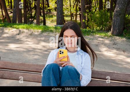 Giovane donna caucasica adulta che si gode una giornata di sole al parco mentre controlla il suo telefono con una custodia gialla su una panchina di legno. Stile di vita digitale nomade. Foto Stock