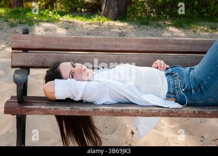 Giovane donna caucasica in un abbigliamento informale che si rilassa su una panchina di legno in una spiaggia sabbiosa in una giornata di sole con la natura sullo sfondo. Il lifest dei nomadi digitali Foto Stock