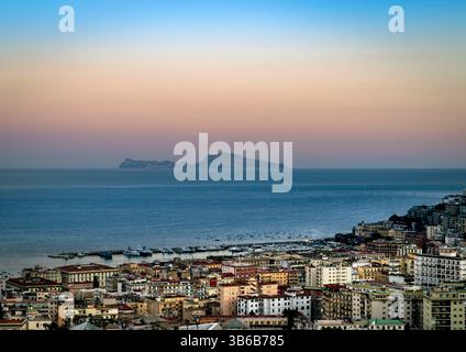 Una vista panoramica di una città costiera al tramonto, caratterizzata da edifici colorati e un mare sereno. Un'isola è visibile in lontananza sotto un cielo in pendenza t Foto Stock