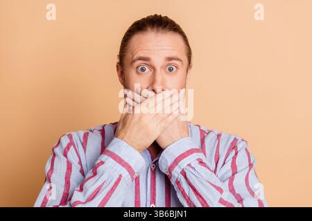 Giovane dal look sorpreso copre la bocca con le mani, indossando una camicia casual a righe davanti al caldo sfondo beige Foto Stock
