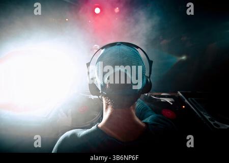 Primo piano del dorso della testa di un DJ maschio sul palco con un cappello da baseball, a un concerto in un nightclub buio Foto Stock