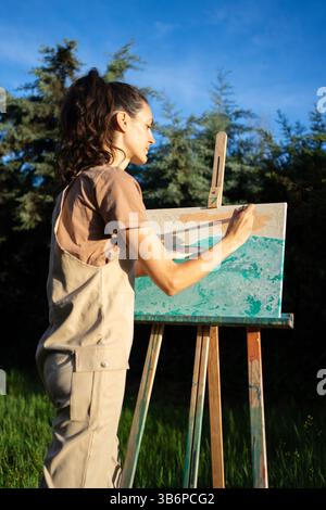 Artista donna che dipinge su tela con pitture ad olio usando pennello e cavalletto durante la creazione artistica plein air in natura Foto Stock