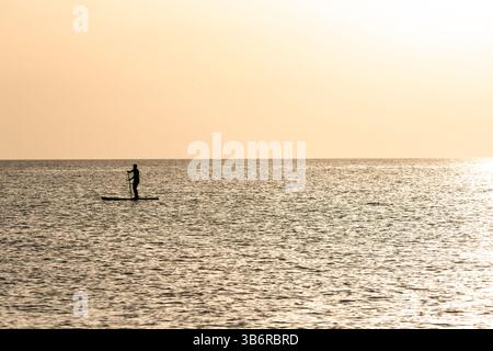Silhouette di una sola persona in paddleboard su un mare calmo durante un tramonto dorato. Ideale per concetti relativi al benessere, alla libertà, all'equilibrio e al natu Foto Stock