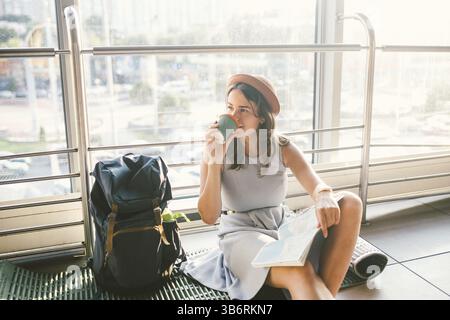 In attesa del trasporto ritardato nel terminale dell'aeroporto o stazione ferroviaria. Giovane donna caucasica nel vestire e hat siede sul tappeto turistica con zaino n Foto Stock