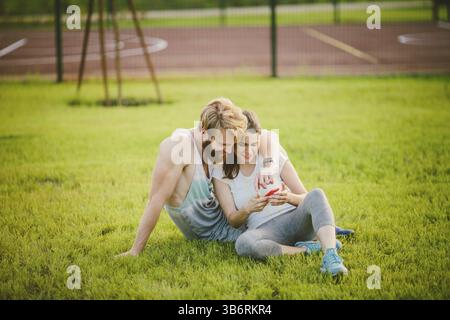 Giovane eterosessuale caucasico con la barba riposata dopo un allenamento all'aperto in un parco sul prato, erba verde seduta in un abbraccio e usando una te Foto Stock