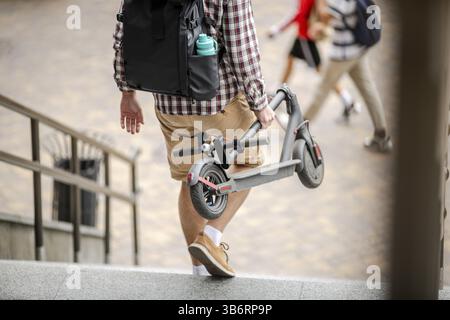 Uomo che trasporta e-Scooter piegato in mano sulle scale vicino all'edificio. Trasporto urbano ecologico, concetto di città moderna. Stile di vita tecnologico ecologico. Foto Stock