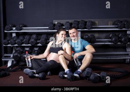 Coppia caucasica uomo forte e muscoloso e donna incinta sportiva che riposa mentre si siede sul pavimento vicino alle attrezzature fitness in palestra per il cross training. SP Foto Stock