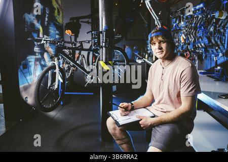 Bel giovane uomo elegante in un cappuccio di snapback e con un tatuaggio proprietario di piccola impresa la vendita di una bicicletta, il workshop si siede sullo sfondo della stor Foto Stock