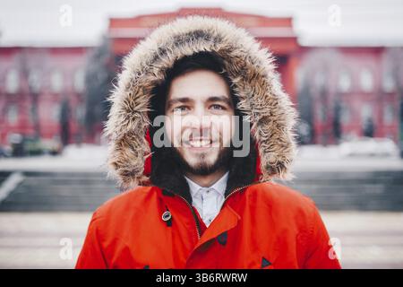 Ritratto, close-up di giovani con elegante uomo sorridente con la barba vestito di rosso giacca invernale con un cappuccio e pelliccia sulla sua testa si erge contro l'backgro Foto Stock