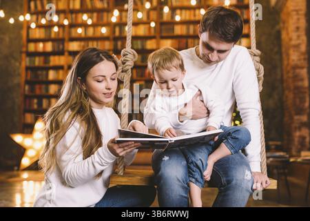 Insieme di famiglia. Mamma, papà e figlio leggono insieme un libro di storie seduti a casa la sera dello swing. Concetto di famiglia e genitorialità. La famiglia felice legge Foto Stock