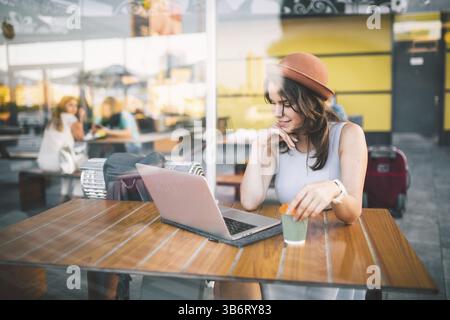 Bella giovane donna caucasica seduta su una terrazza in un bar in estate ad un tavolo di legno con un cappello e un piatto utilizza la tecnologia, lavorando dietro un lapt Foto Stock