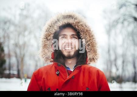 Ritratto, close-up di giovani con elegante uomo sorridente con la barba vestito di rosso giacca invernale con un cappuccio e pelliccia sulla sua testa si erge contro l'backgro Foto Stock