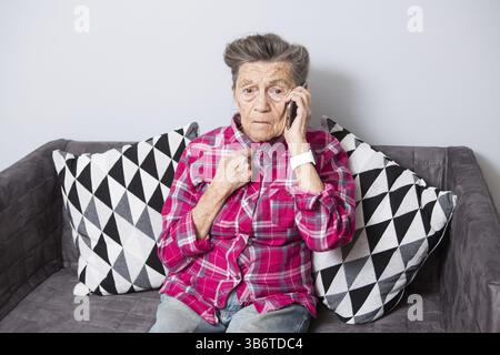 Una vecchia nonna anziana con i capelli grigi si siede a casa sul divano usando il telefono, una conversazione telefonica per sentire le cattive notizie. Emozione Foto Stock