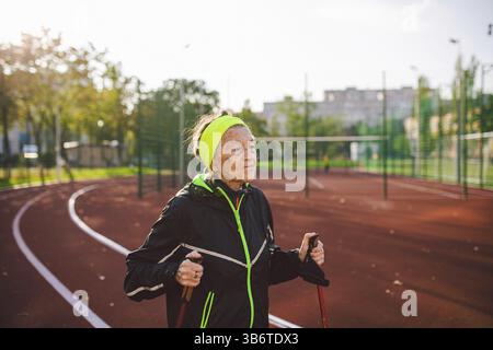 Donna anziana che cammina con i bastoncini nello stadio su una copertura di gomma rossa. Donna anziana 88 anni che fa esercizi di nordic walking presso la città di stadiu Foto Stock