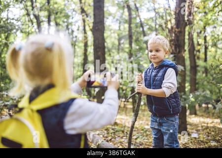 Bambini I bambini in età prescolare Caucasian fratello e sorella di scattare foto di ogni altro sul telefono cellulare fotocamera in Forest park autunno. tema di hobby e attivo Foto Stock