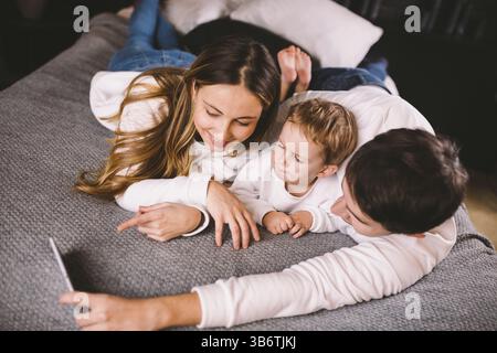 Una giovane famiglia giace sul letto e guarda il telefono cellulare. Mamma, papà e figlio guardano un video su uno smartphone in camera da letto la sera. FAM Foto Stock