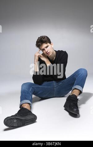 Elegante ragazzo che posa in studio seduto sul pavimento con pantaloni in denim e felpa nera. Ritratto studente maschio completo su sfondo bianco. Studio ha sparato ad Han Foto Stock