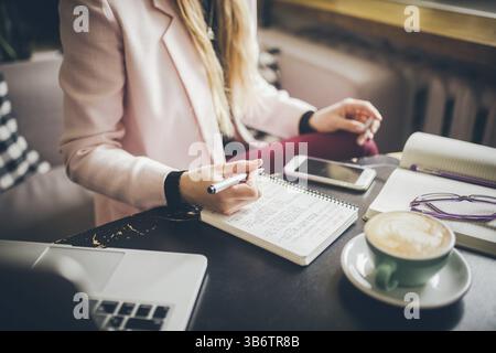 Primo piano donna caucasica mano su un tavolo di legno all'interno del caffè prende appunti sul taccuino. Soggetto libero professionista blogger giornalista al lavoro. Perso irriconoscibile Foto Stock