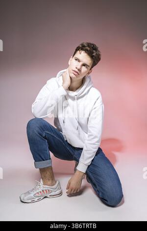 Pensivo ragazzo che posa seduto sul pavimento nello studio. ritratto di uno studente vestito con stile. Studio ha girato un bell'uomo europeo sittin Foto Stock