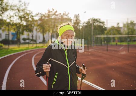 Donna anziana che cammina con i bastoncini nello stadio su una copertura di gomma rossa. Donna anziana 88 anni che fa esercizi di nordic walking presso la città di stadiu Foto Stock