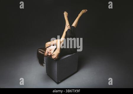 Tema sociale. Giovane donna caucasica in strana posa in abito in posa su cubo nero, sfondo scuro, simboleggia dolore, sofferenza, cercare aiuto, protecti Foto Stock