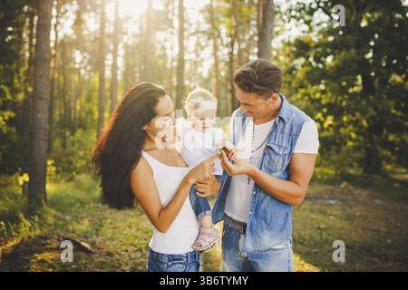 Elegante e giovane famiglia, mamma, papà e figlia bionda di un anno seduto con il padre sulle spalle che giocano felici e sorridenti, fuori dalla città i Foto Stock