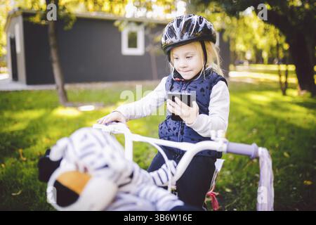 Funny bambino Caucasian ragazza bionda in una bicicletta Casco nei pressi di un viola moto con un cestello esterno al parco su un prato verde erba carrello a casa. Gioca u Foto Stock