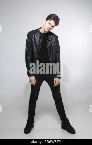 Giovane ragazzo caucasico che posa in studio, ritratto a figura intera di un modello in abiti neri. Bella adolescente in giacca nera, jeans e sneakers su un g Foto Stock