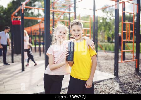 I bambini caucasici fratello e sorella si levano in abbraccio a terra di sport all'aperto con sorriso e fiasca con acqua dopo l'allenamento. Gemelli ragazzo e ragazza ath Foto Stock