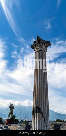 Un'alta e antica colonna con un capitello decorativo si erge su un cielo blu brillante con nuvole mostruose. Sullo sfondo è visibile una statua di una figura Foto Stock