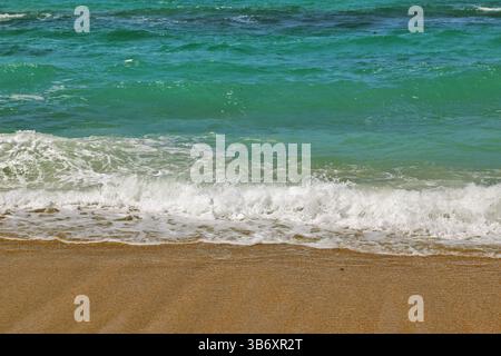 Le limpide onde turchesi rotolano dolcemente su una spiaggia di sabbia dorata sotto la luce del sole, catturando una tranquilla scena costiera ideale per viaggi e temi naturalistici. Foto Stock