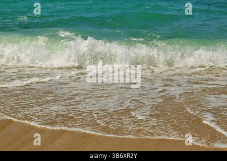 Le limpide onde turchesi rotolano dolcemente su una spiaggia di sabbia dorata sotto la luce del sole, catturando una tranquilla scena costiera ideale per viaggi e temi naturalistici. Foto Stock