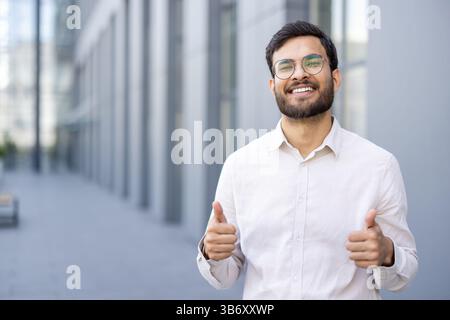 Un allegro uomo d'affari mostra un gesto con il pollice in alto, in piedi all'aperto con un sorriso brillante, celebrando il successo o l'approvazione. Foto Stock