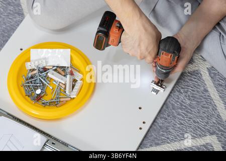 Primo piano di mani mans utilizzando un trapano elettrico per avvitare insieme le parti durante l'assemblaggio di un armadietto a casa. Foto Stock