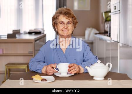 Donna anziana felice che si gusta il tè a casa, sorride mentre tiene la tazza, seduto al tavolo della cucina con teiera e dolci in un ambiente interno accogliente e moderno Foto Stock